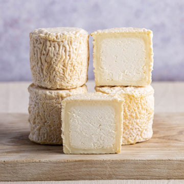 Whole and cut pieces of petit blaja goat's cheese on a wooden chopping board.