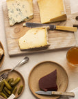 Cuts of cheese on a wooden board, next to plates of cornichons, quince, bread and a glass of wine.