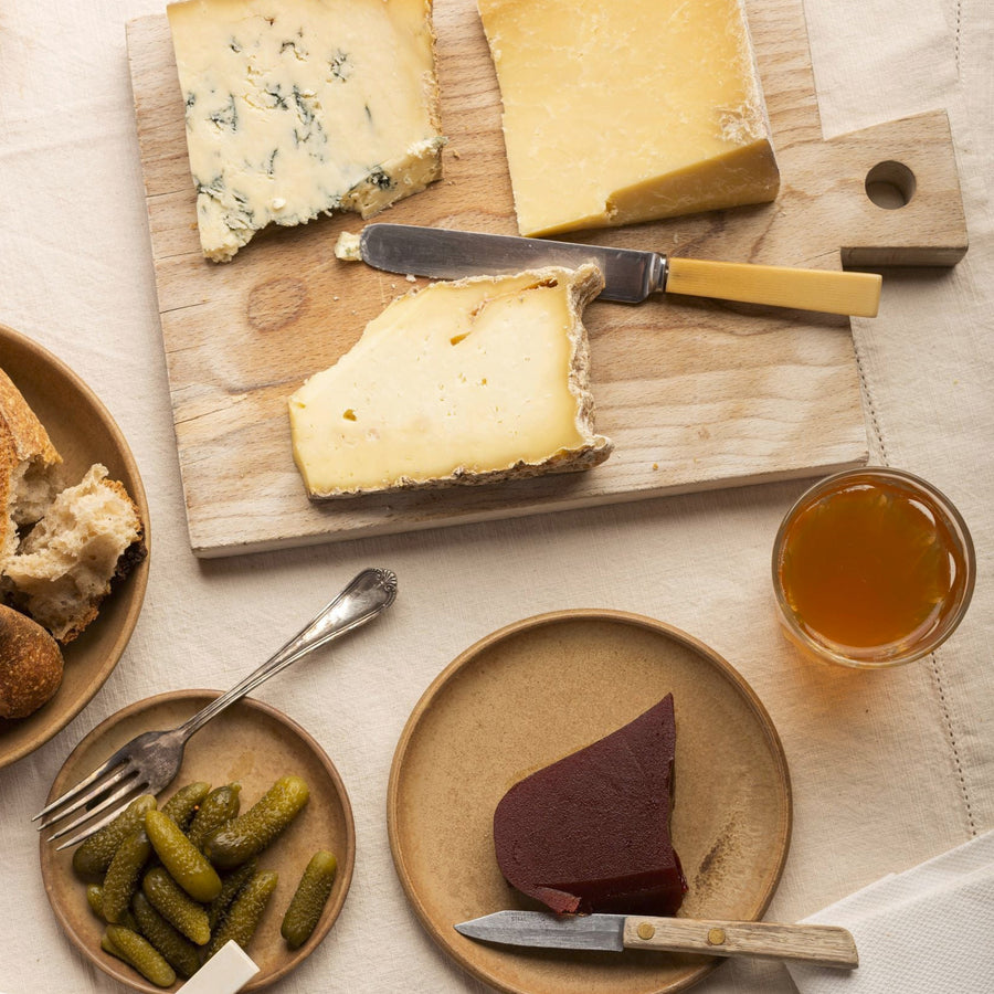 Cuts of cheese on a wooden board, next to plates of cornichons, quince, bread and a glass of wine.