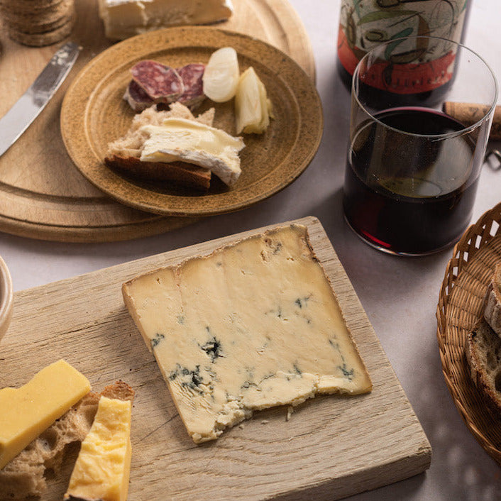 A piece of Stilton blue cheese on a wooden board, next to a plate with bread and cheese, and glass of red wine.