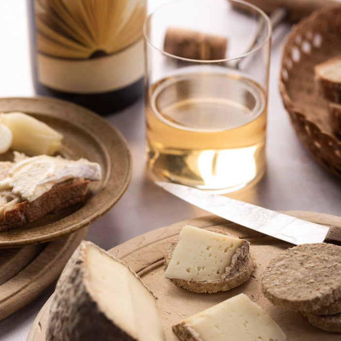 Cuts of Pecorino cheese and a knife on a wooden board, with a glass of white wine in the background.