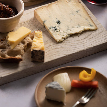 Pieces of cheese on a wooden board, with a plate of pickles in the foreground.