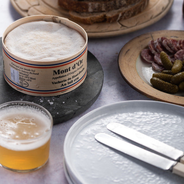 A plate with some cutlery, a glass of beer and a whole Vacherin cheese on a slate platter.