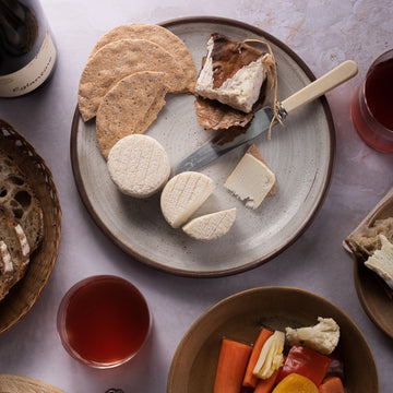 Cheese and biscuits on a plate, next to a glass of red wine and a plate of pickles. 