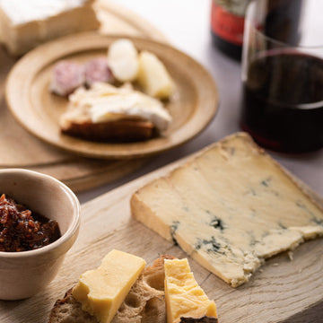 A slice of Stilton blue cheese on a wooden board next to some Cheddar cheese on a slice of bread, with plates of accompaniments.
