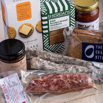 A selection of biscuits for cheese, salamis and jars of pickle on a wooden board.