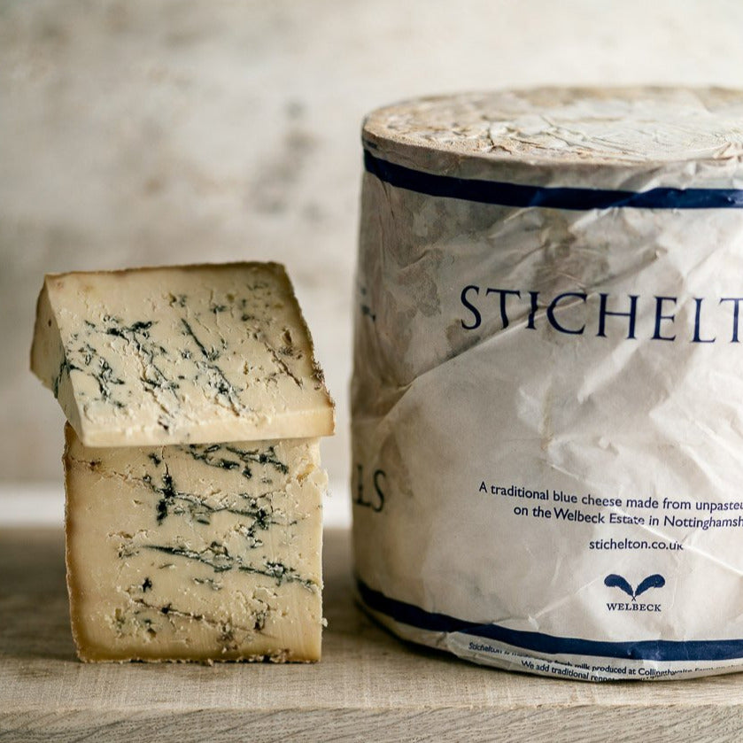 Two pieces of cut Stichelton blue cheese on a board next to a whole, wrapped cheese.