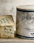 Two pieces of cut Stichelton blue cheese on a board next to a whole, wrapped cheese.